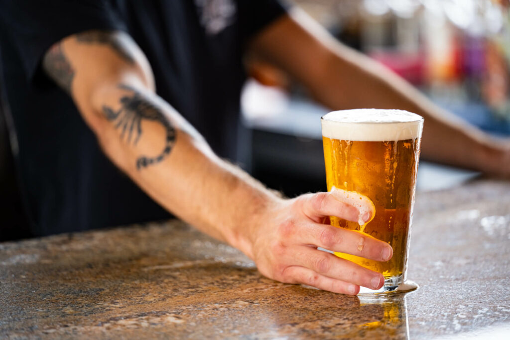 A person with a scorpion tattoo on their arm places a pint of beer with a frothy head on a granite countertop.