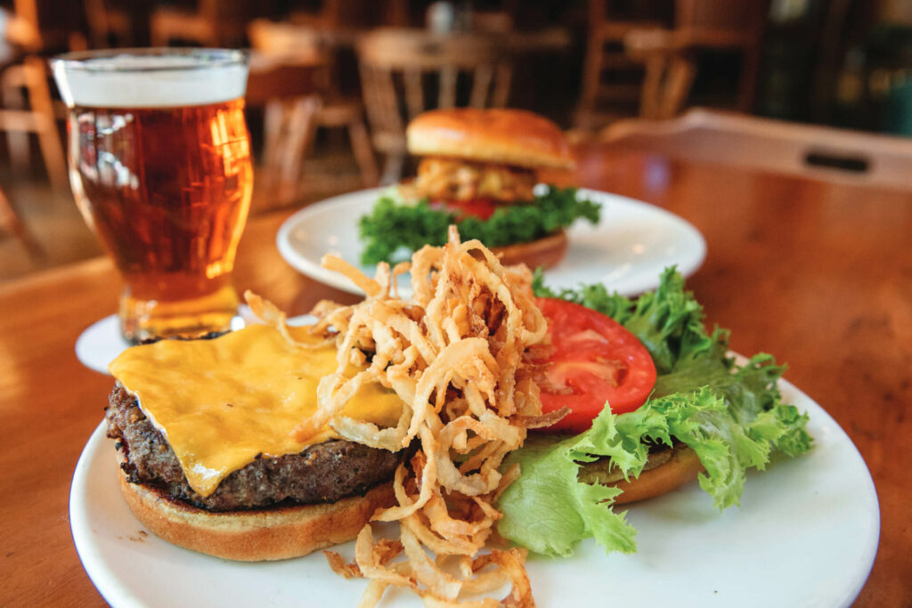 A close-up of a cheeseburger topped with crispy fried onions, lettuce, and tomato on a white plate, next to a glass of beer; another burger is blurred in the background.